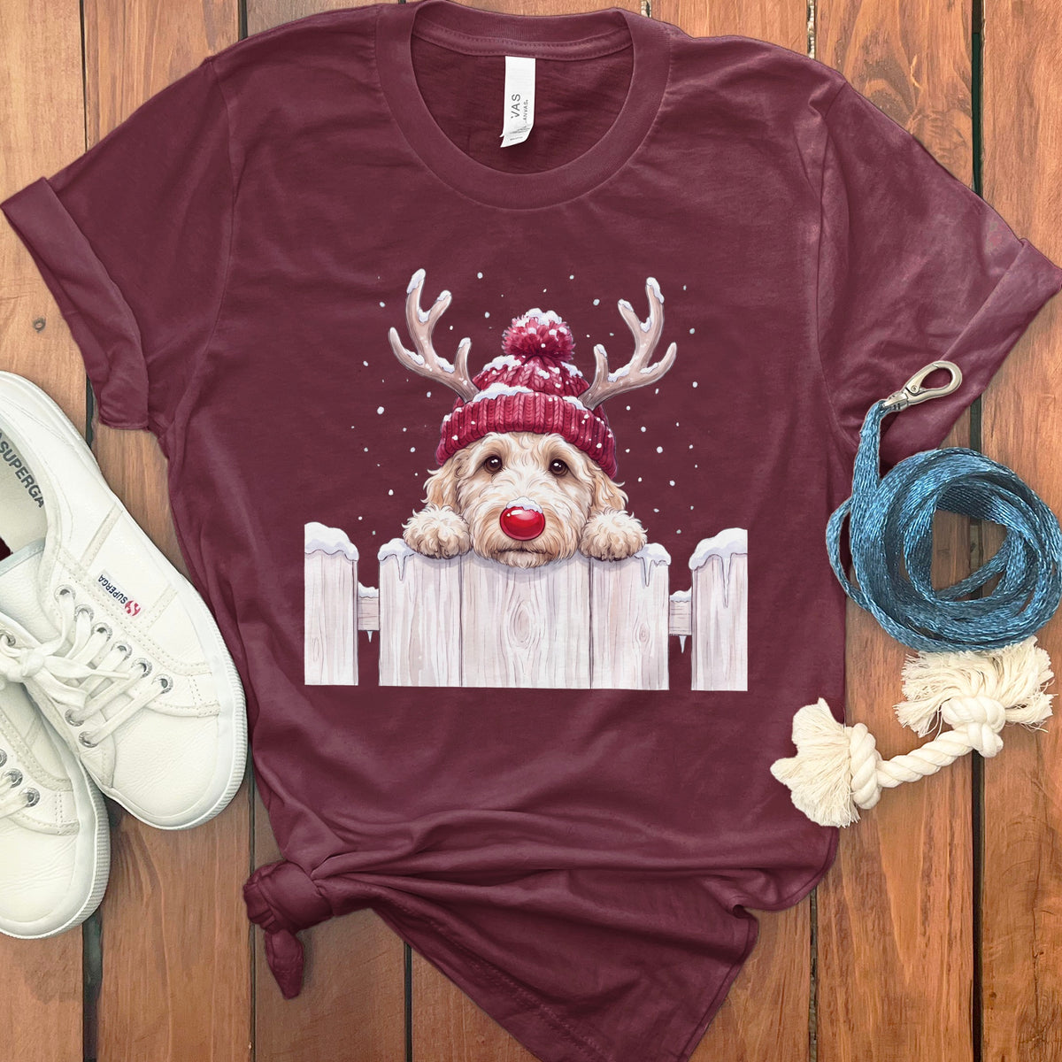 Maroon t-shirt featuring a doodle dog with antlers and a red nose peeking over a snowy fence.