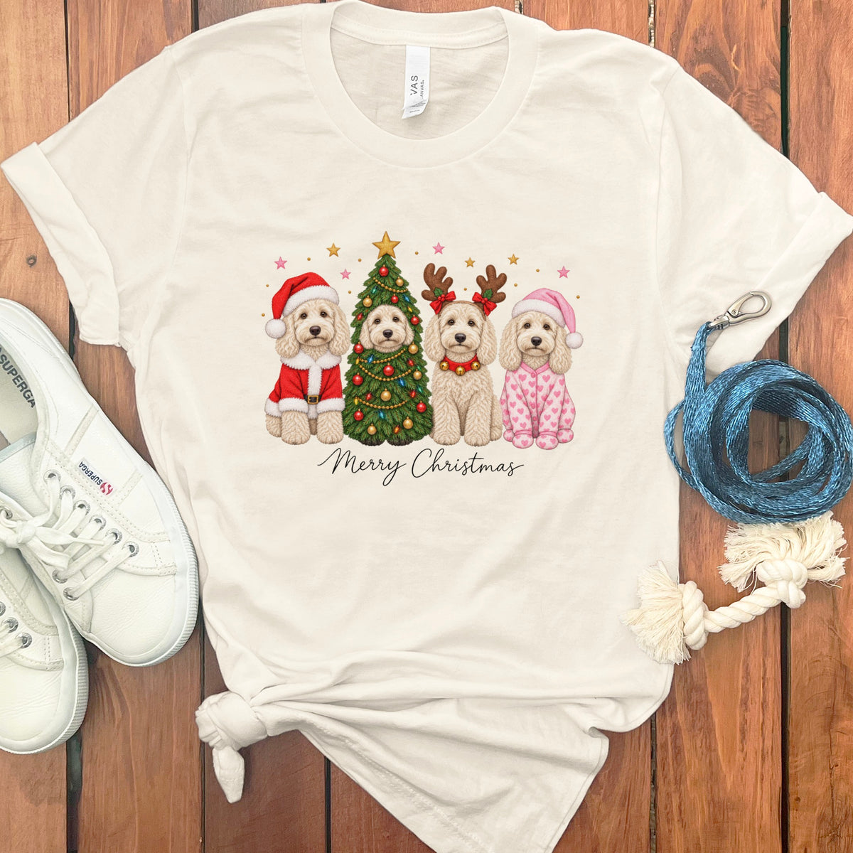 A cream-colored t-shirt with a graphic of four doodle dogs celebrating Christmas, surrounded by white sneakers and accessories on a wooden background.