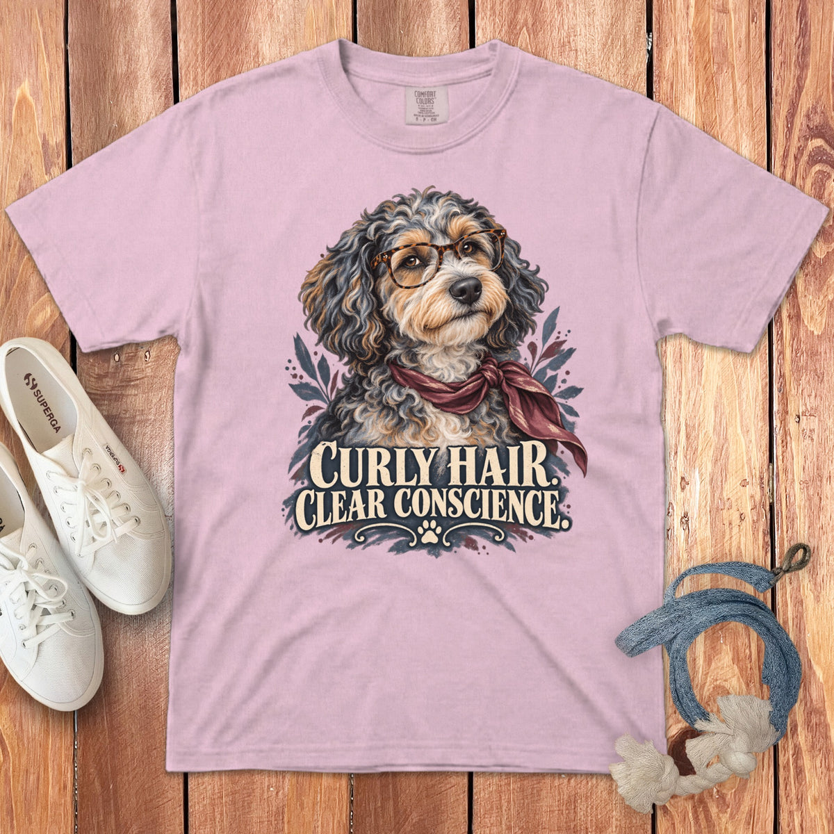 A light pink t-shirt with a graphic of a curly-haired dog wearing glasses and a scarf, accompanied by the text Curly Hair Clear Conscience.