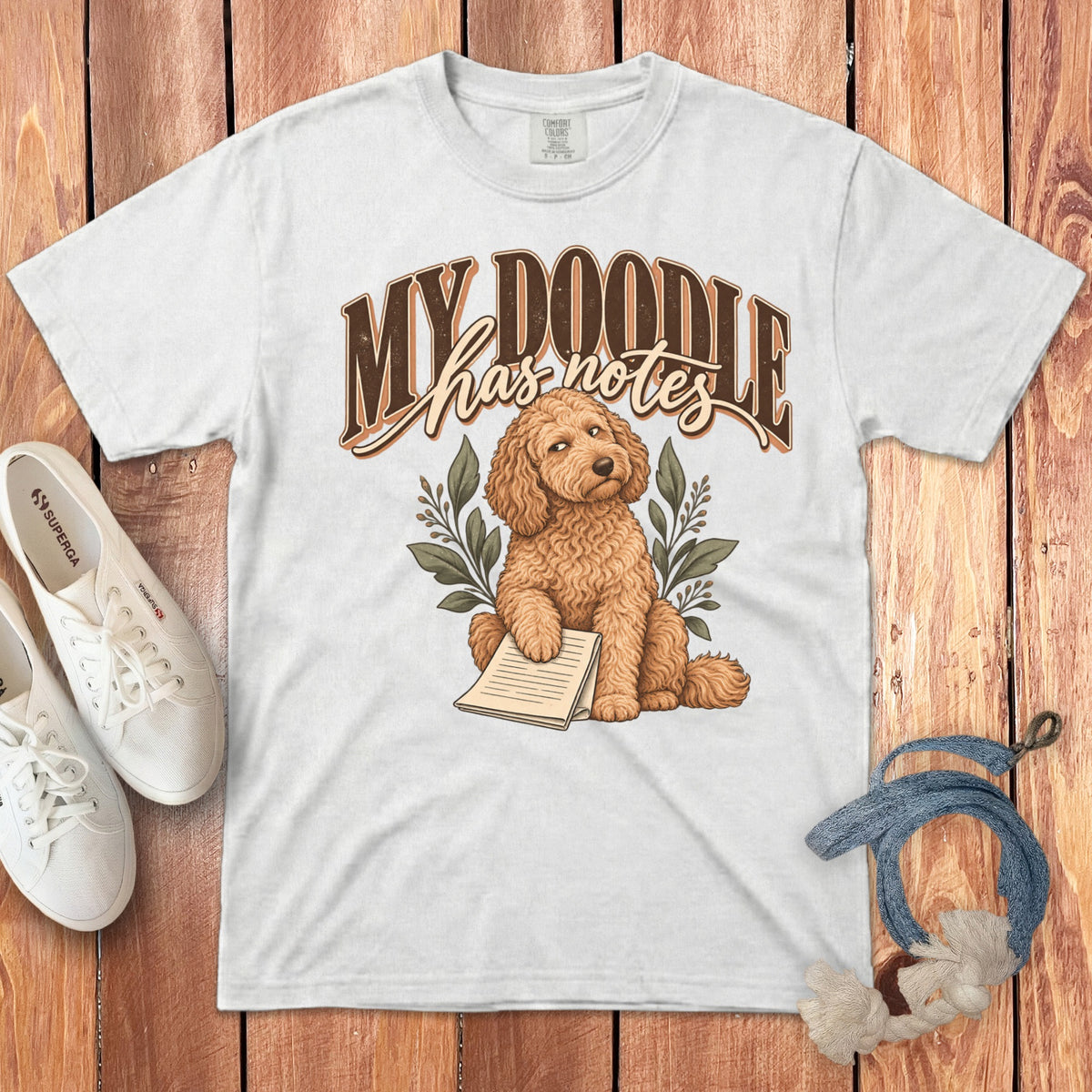 A white t-shirt with a graphic of a curly-haired doodle dog holding a notepad, accompanied by leaves and text that reads "My Doodle has notes."