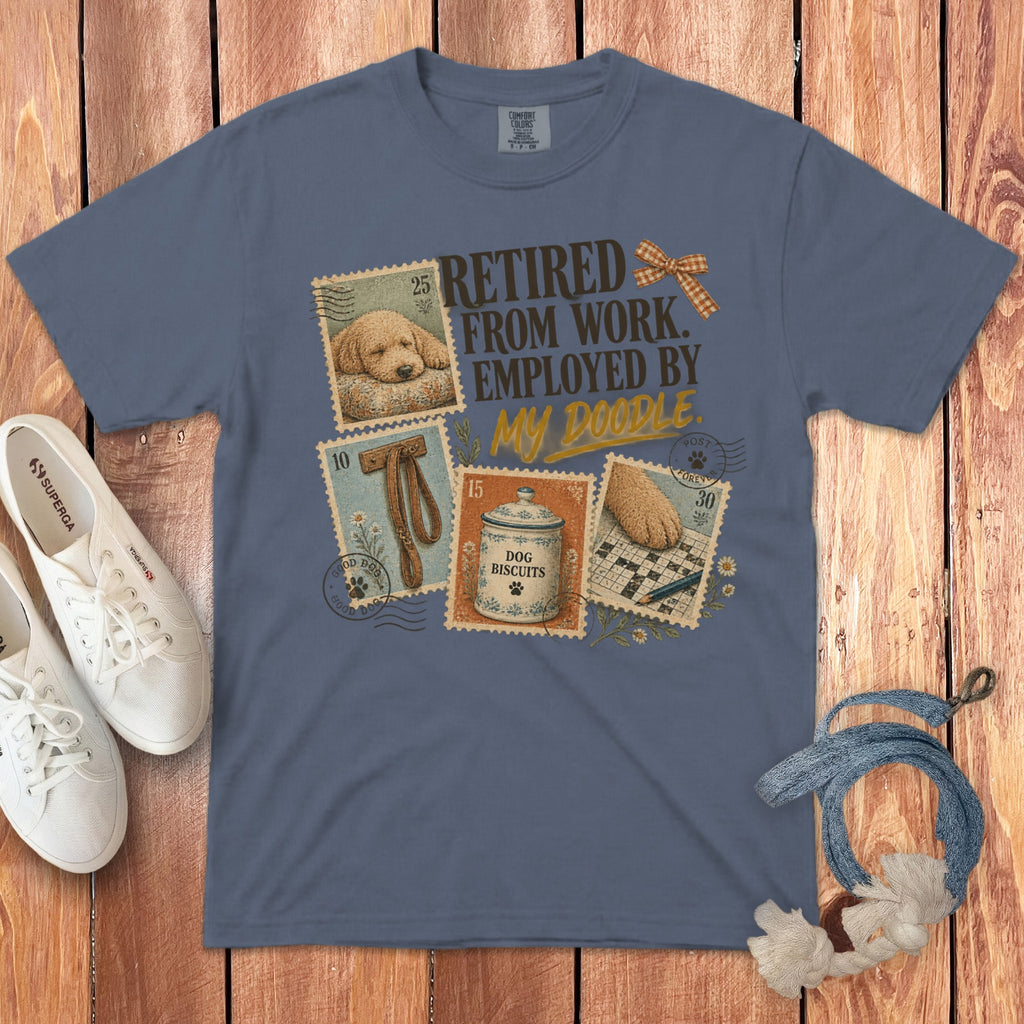 A blue t-shirt with a graphic of postage stamps and text reading Retired From Work Employed By My Doodle, laid flat on a wooden surface with white sneakers and a dog leash.