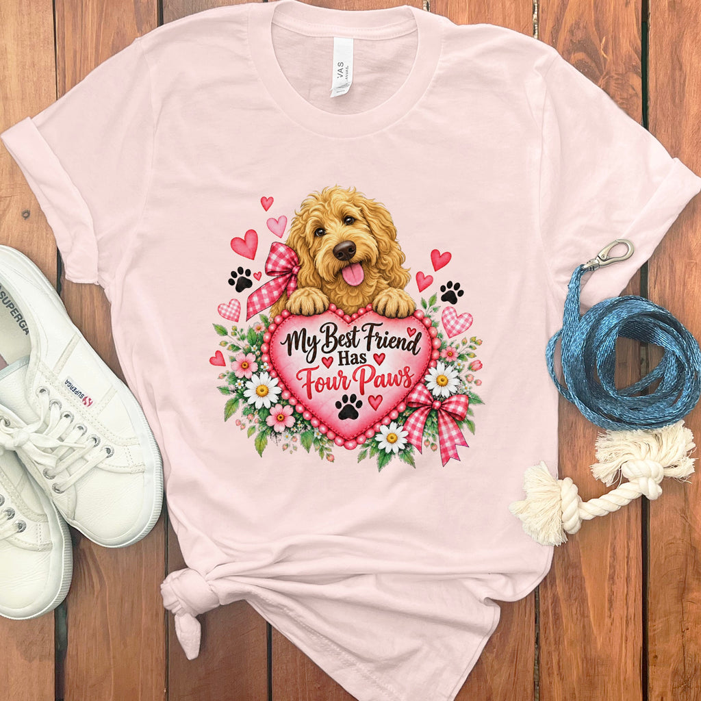 A light pink t-shirt with a graphic of a golden doodle puppy holding a pink heart that says My Best Friend Has Four Paws, surrounded by flowers, paw prints, and small hearts.