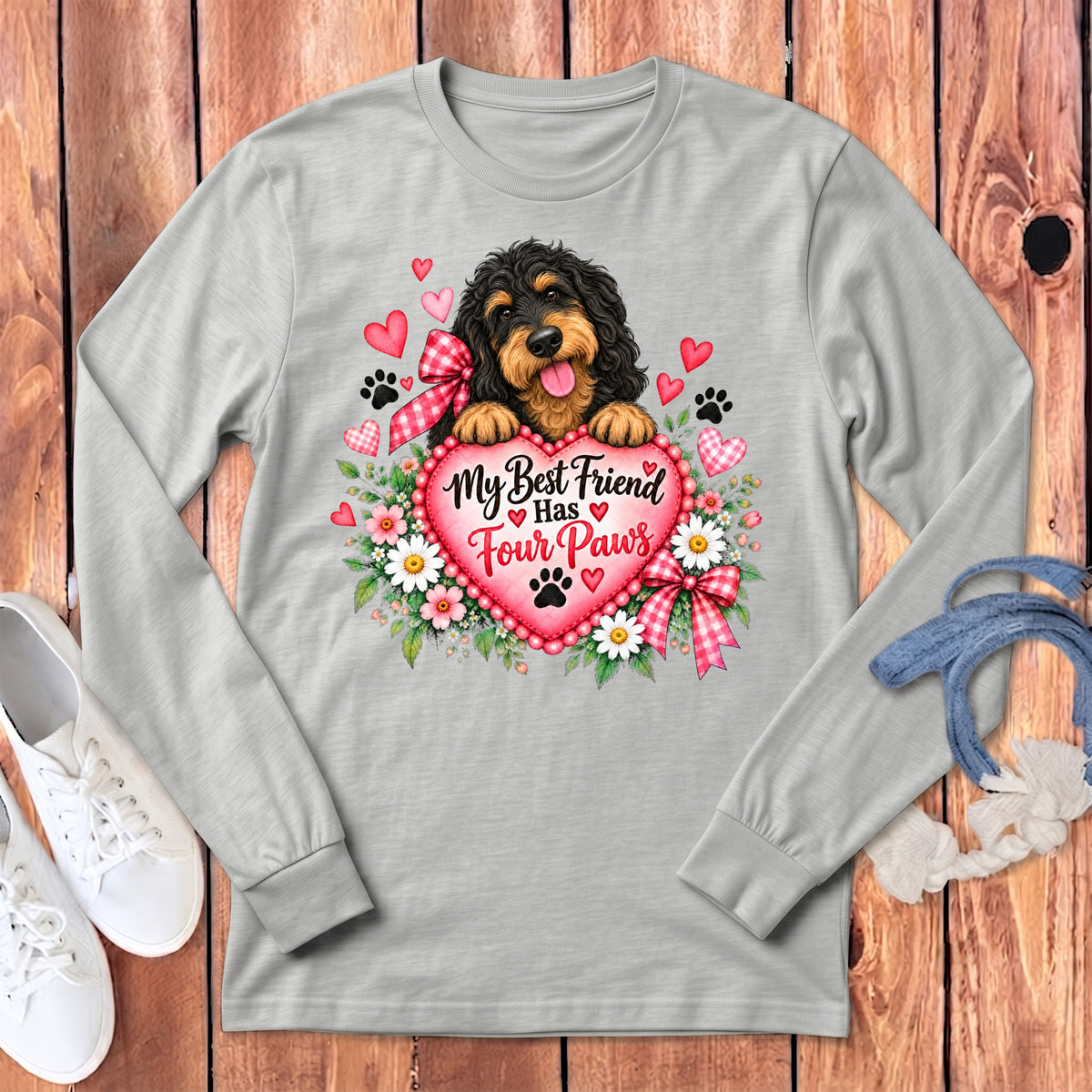 A light gray long-sleeve t-shirt featuring a doodle dog holding a heart with text that reads My Best Friend Has Four Paws, surrounded by flowers, hearts, and paw prints, laid flat on a wooden surface with white sneakers.