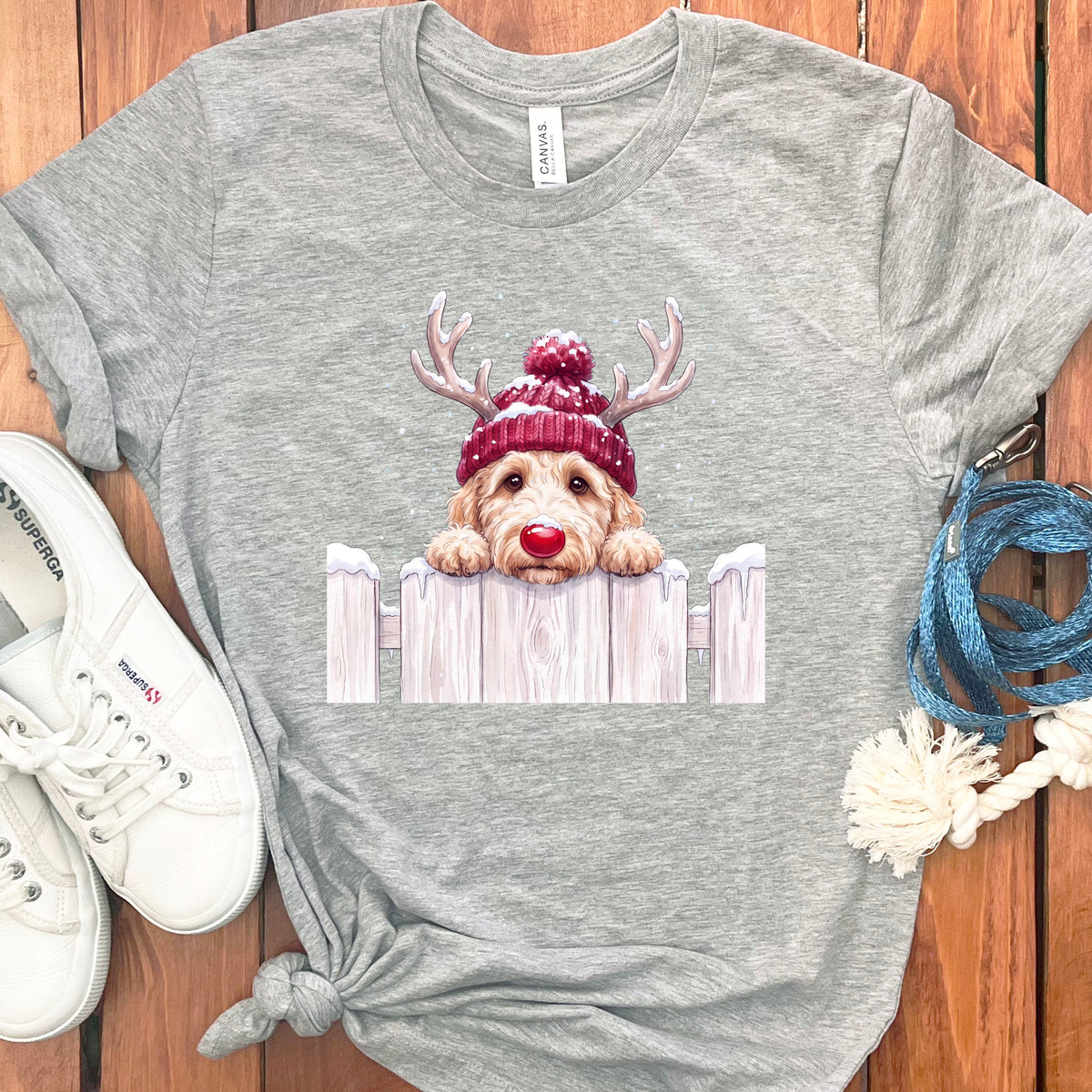 Light grey t-shirt with a goldendoodle wearing a red beanie and antlers peeking over a fence.