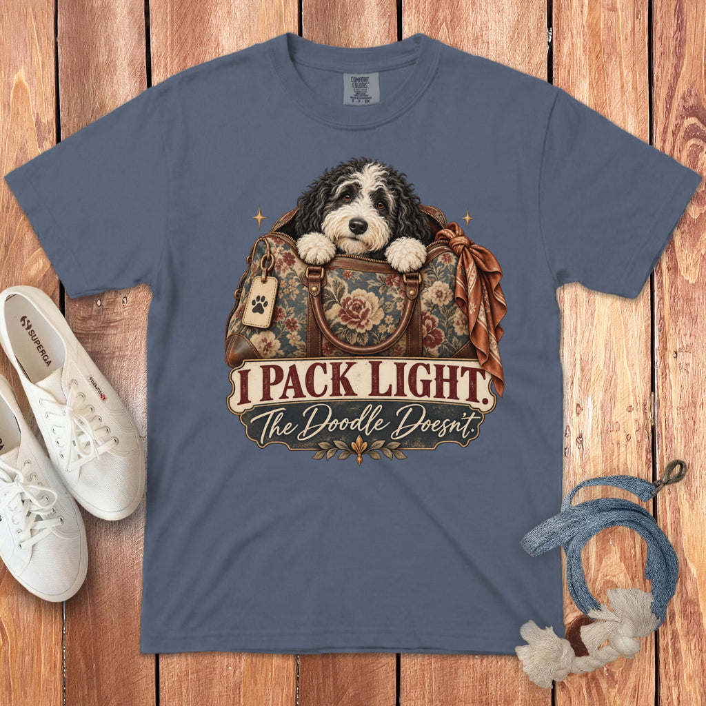 A slate blue t-shirt featuring an illustration of a doodle puppy in a floral bag with text reading I Pack Light The Doodle Doesnt.