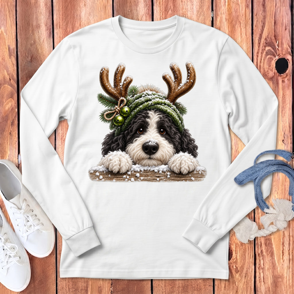Bernedoodle dog wearing Santa hat and antlers, peeking over a snowy log.