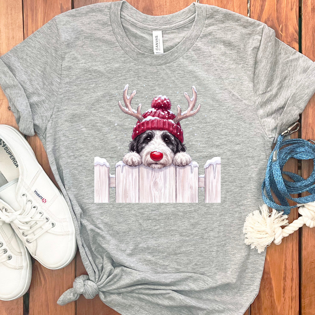 Grey t-shirt with a doodle of a dog wearing a red beanie, antlers, and red nose peeking over a fence.