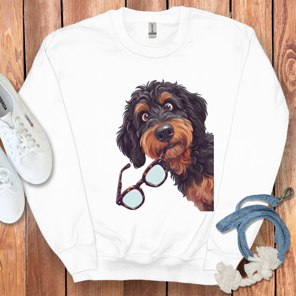 A white sweatshirt on a wooden background features a graphic of a black and brown doodle dog holding glasses in its mouth.