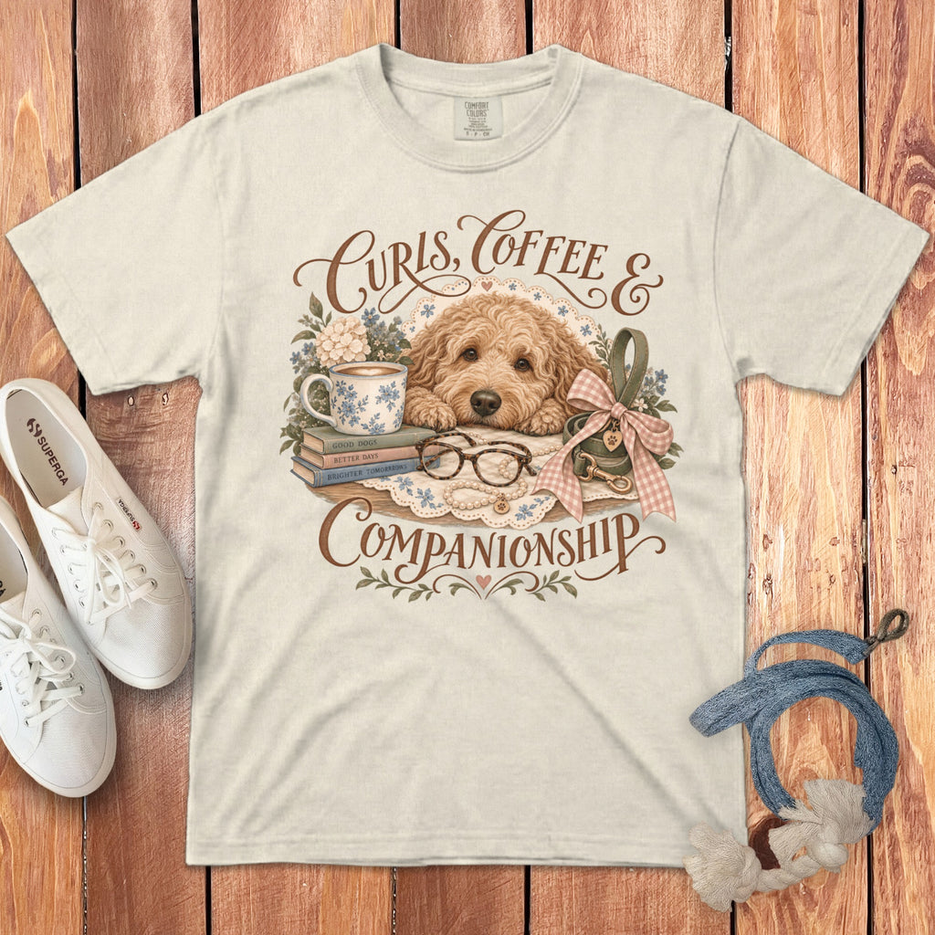 A light-colored t-shirt featuring a graphic of a labradoodle resting near books, coffee, and text reading Curls Coffee and Companionship.