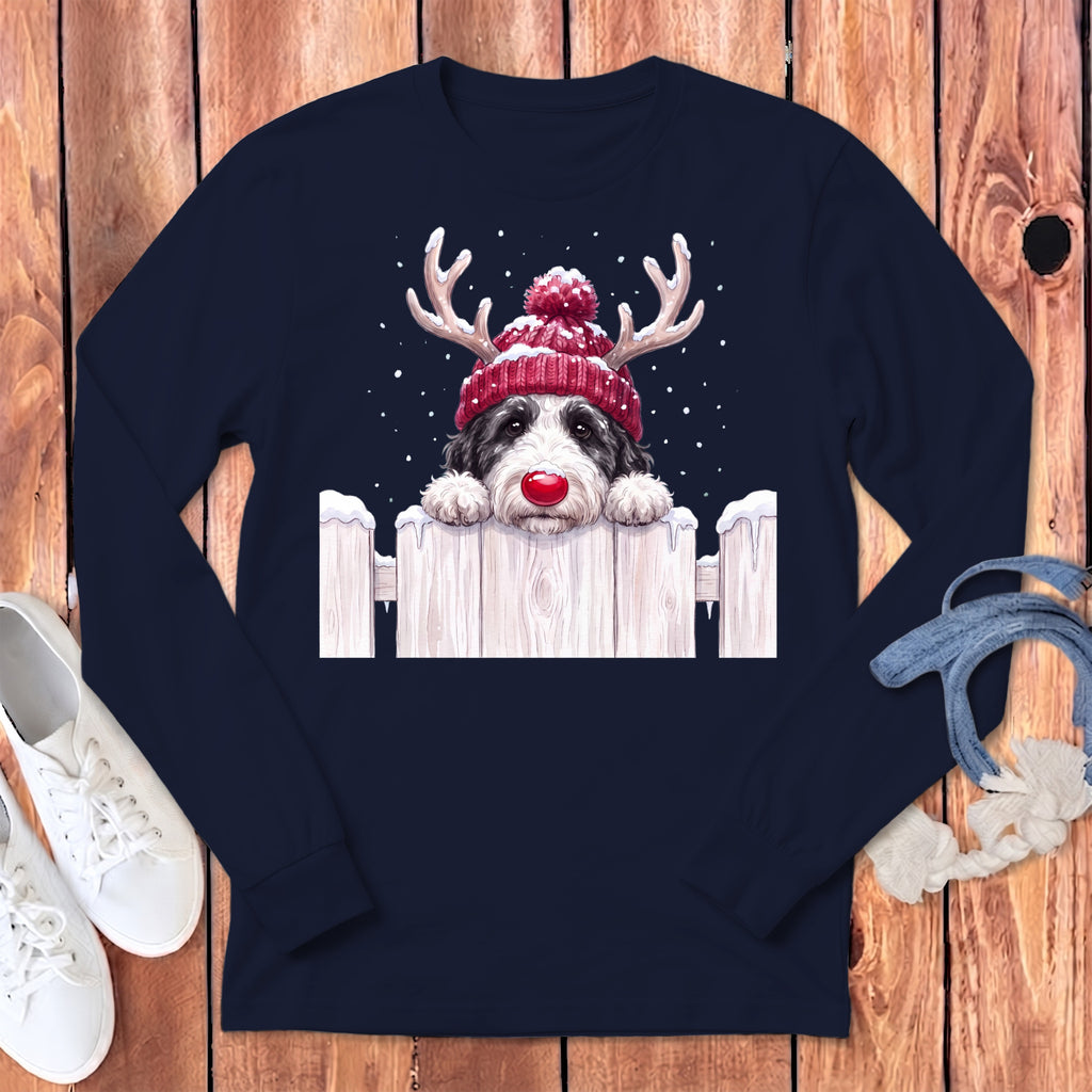Dog with reindeer antlers and red nose peeking over a snow-covered fence.