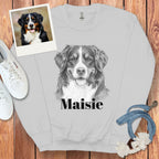 Gray sweatshirt with a dog illustration and 'Maisie' text, placed on a wooden surface with a photo of a dog and pet accessories.
