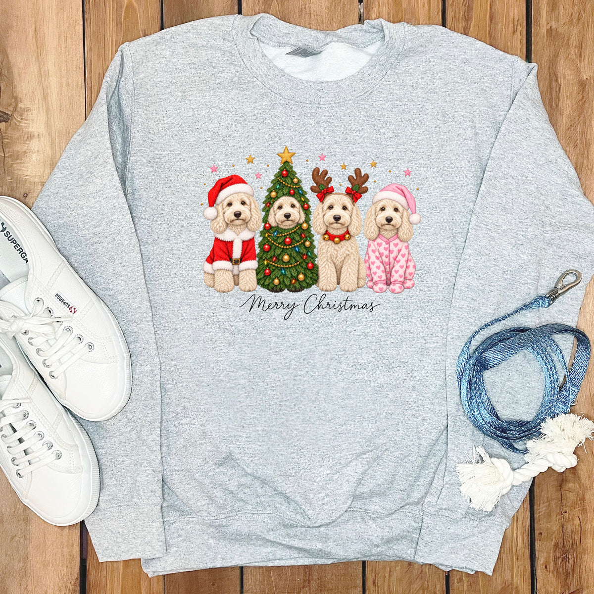 A light gray sweatshirt features a graphic of four doodle dogs celebrating Christmas, surrounded by white sneakers and a blue leash on a wooden background.
