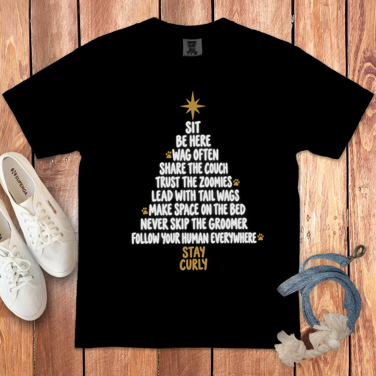 A black t-shirt printed with white and gold text arranged in a Christmas tree shape, accompanied by white sneakers and a dog leash on a wooden background.