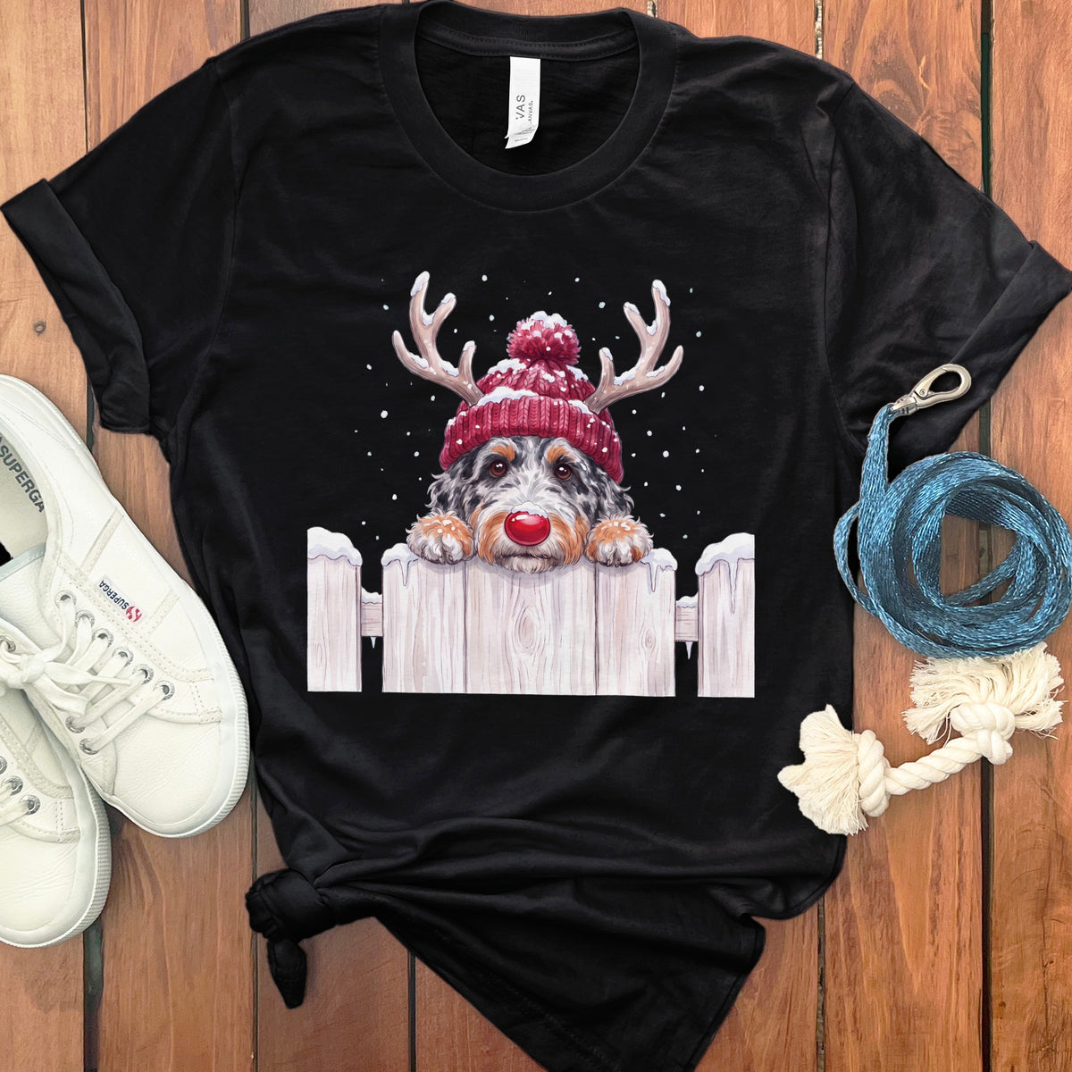 Black t-shirt with a dog wearing a reindeer hat and red nose peeking over a snowy fence.