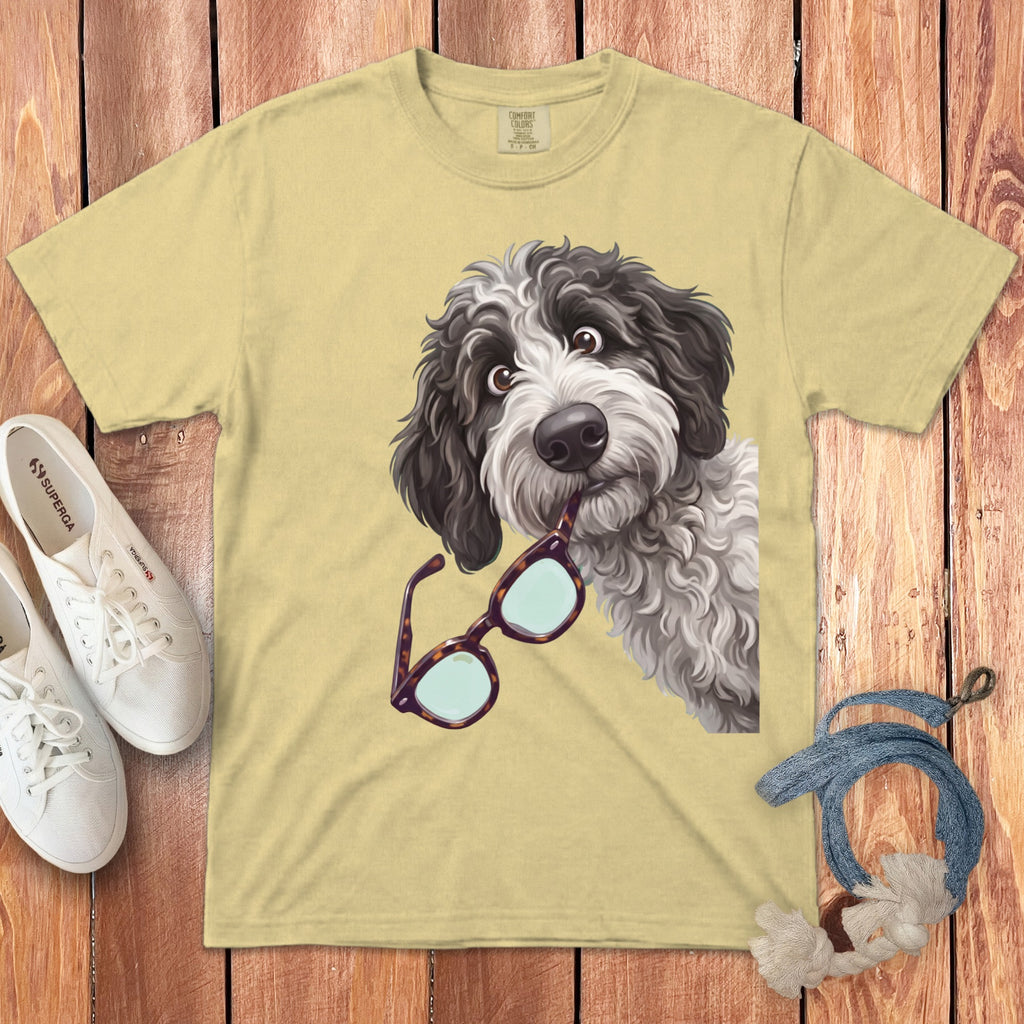 Light yellow t-shirt featuring a cartoon dog holding glasses in its mouth, laid on wood.