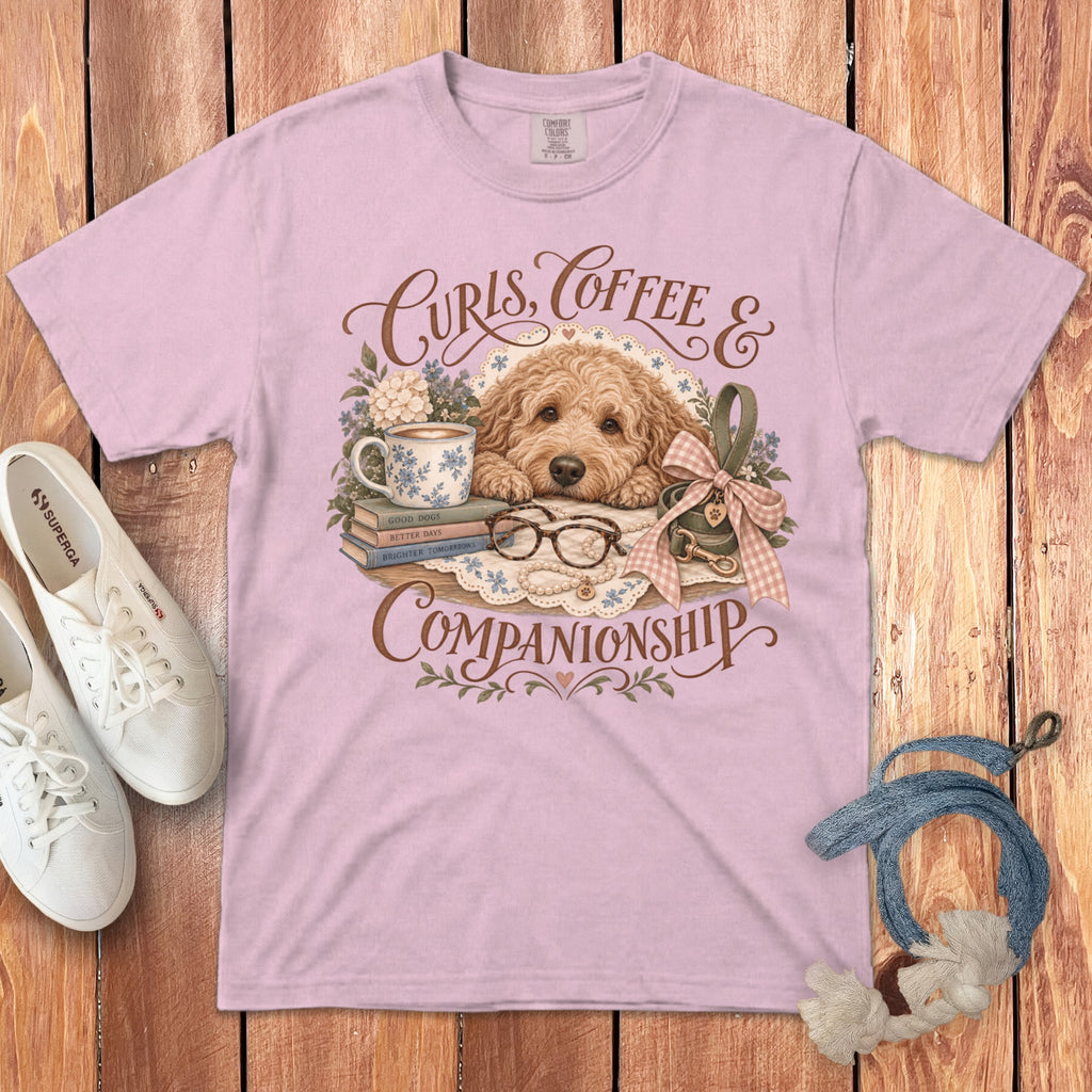 A pink t-shirt featuring a graphic of a Goldendoodle, coffee, books, and the text Curls, Coffee & Companionship.