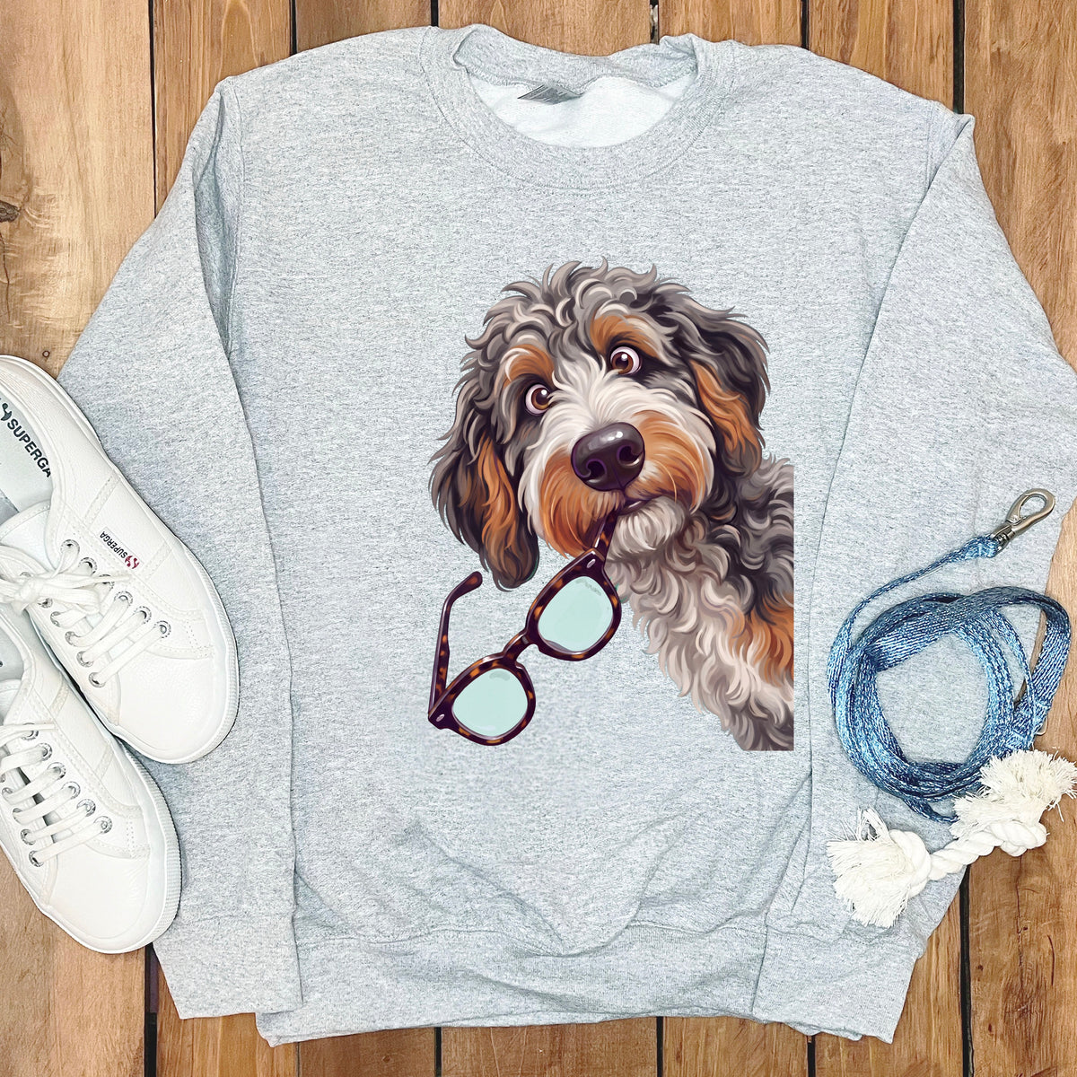 A grey crewneck sweatshirt featuring a graphic of a shaggy dog holding glasses in its mouth, surrounded by white sneakers and a blue leash on a wooden background.