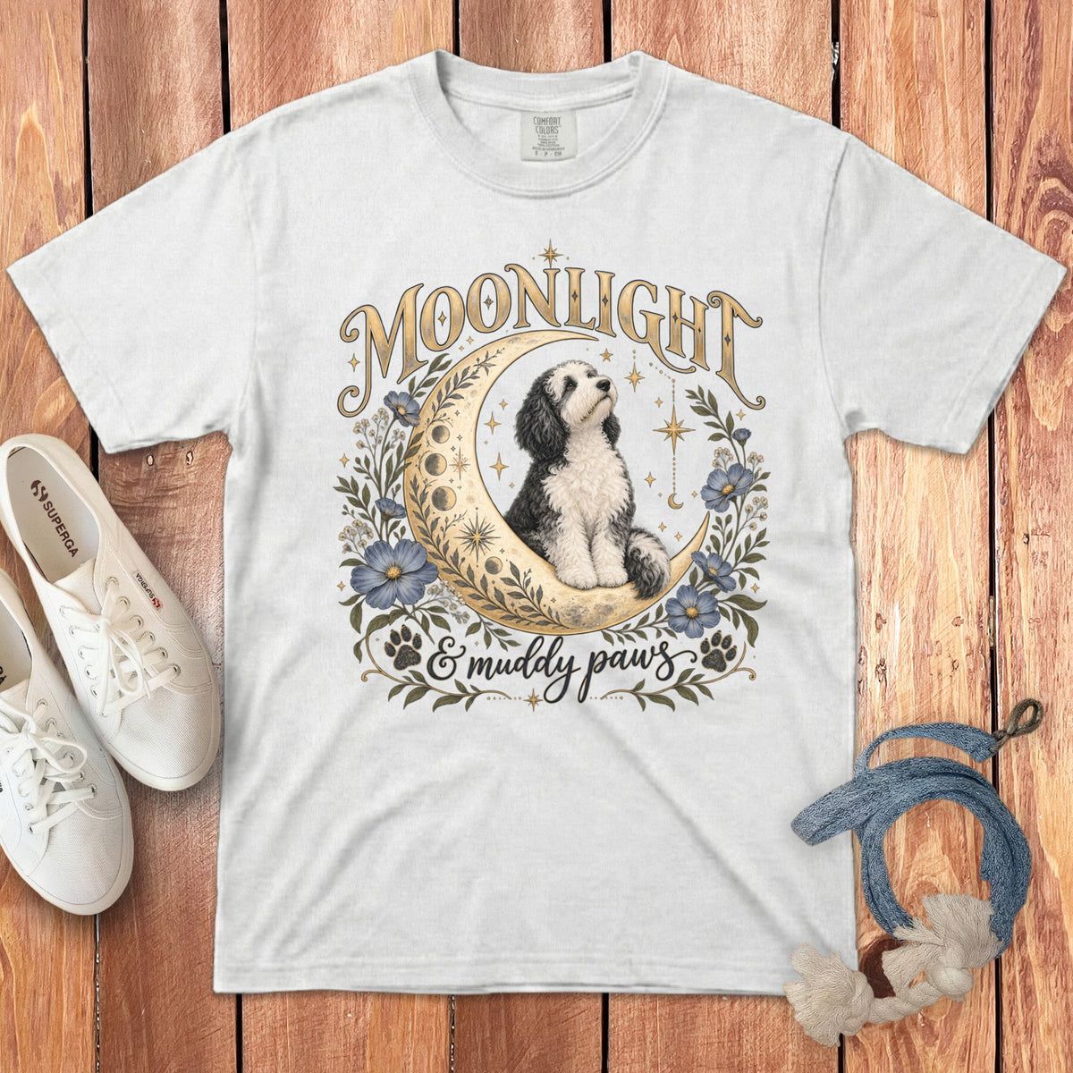 A white crewneck t-shirt featuring a graphic of a dog sitting in a crescent moon with text reading Moonlight & muddy paws, laid flat on a wooden surface with casual shoes and a rope toy.