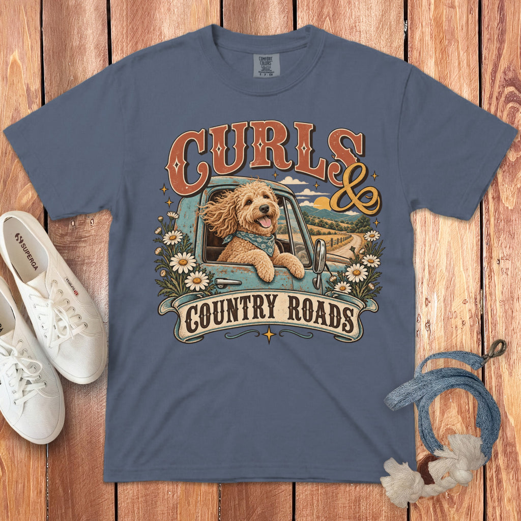 A slate blue t-shirt featuring a graphic of a curly-haired dog in a vintage truck with daisies, accompanied by the text "Curls and Country Roads," laid flat on a wooden surface with white sneakers and a blue rope.