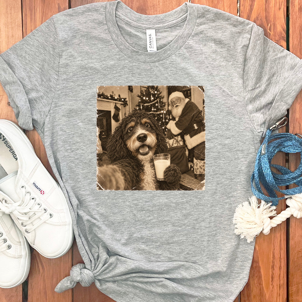 A gray crewneck t-shirt featuring a vintage sepia graphic of a dog holding a glass of milk with Santa Claus in the background, accompanied by white sneakers and a blue leash on a wooden surface.