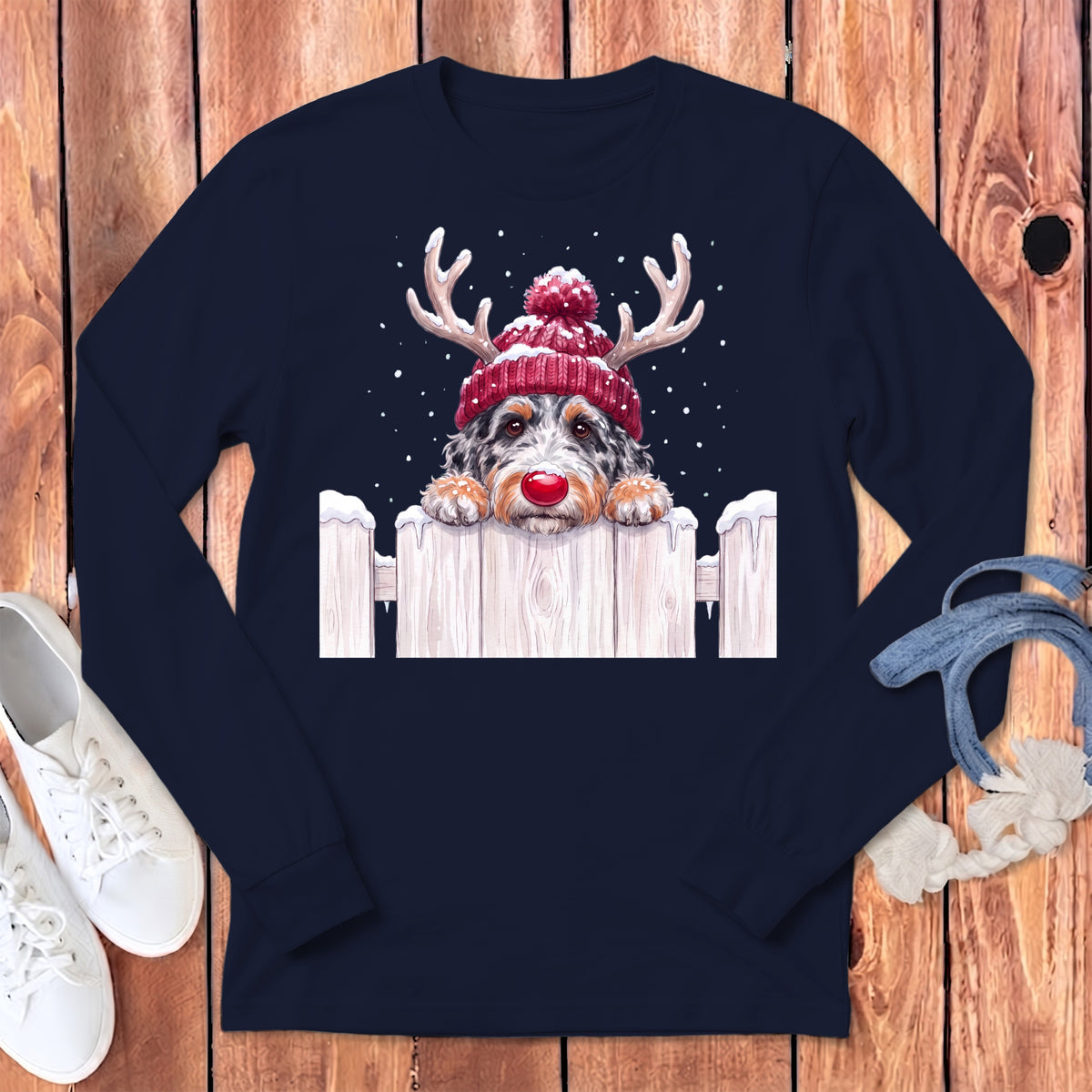 Dog wearing red beanie and antlers peeking over white fence in snow.