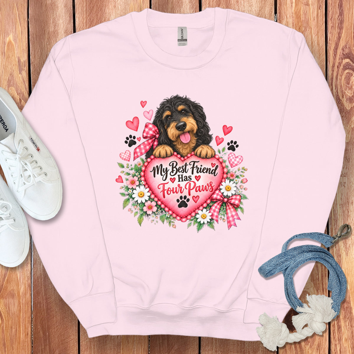 A light pink crewneck sweatshirt features a graphic of a black and tan dog behind a heart with the text My Best Friend Has Four Paws, surrounded by flowers, hearts, and paw prints, displayed on a wooden background with white sneakers and a blue leash.