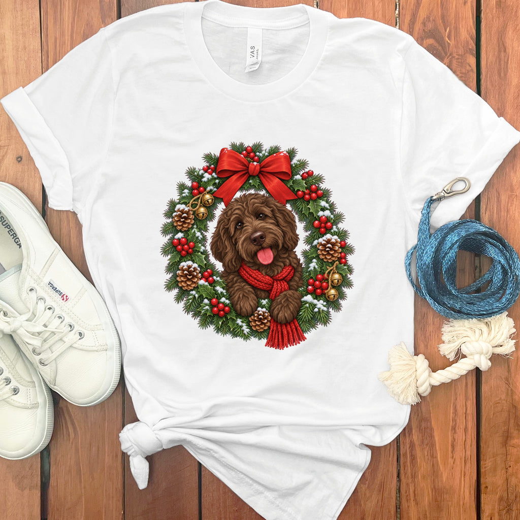 A white t-shirt featuring a graphic of a brown dog wearing a red scarf inside a Christmas wreath, laid on a wooden surface with white sneakers and a blue leash.