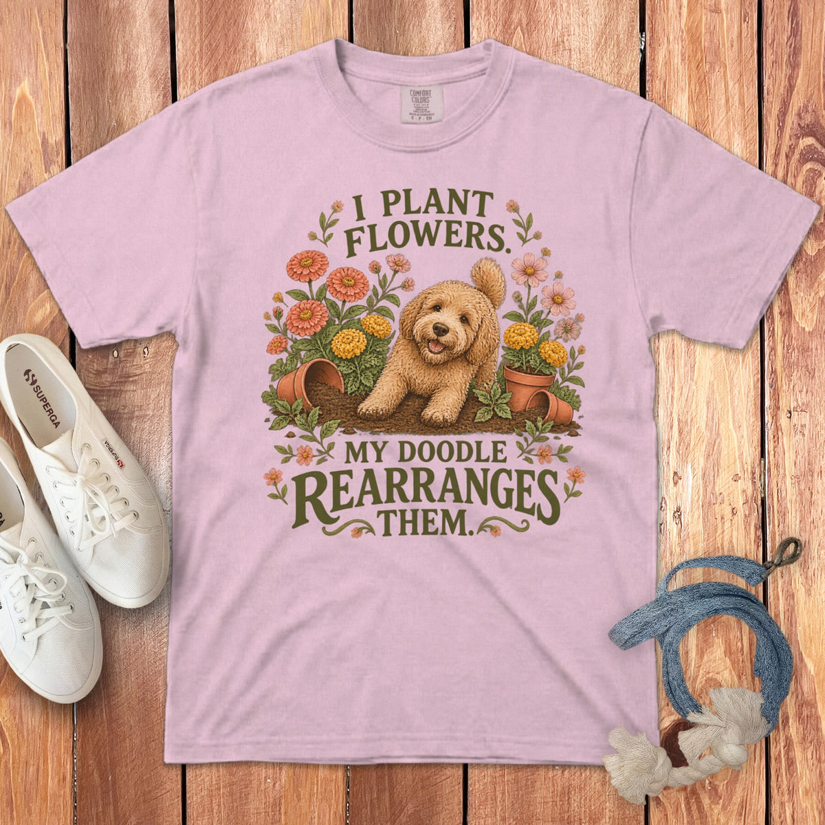 A pink t-shirt featuring a goldendoodle graphic and text that reads I PLANT FLOWERS MY DOODLE REARRANGES THEM.