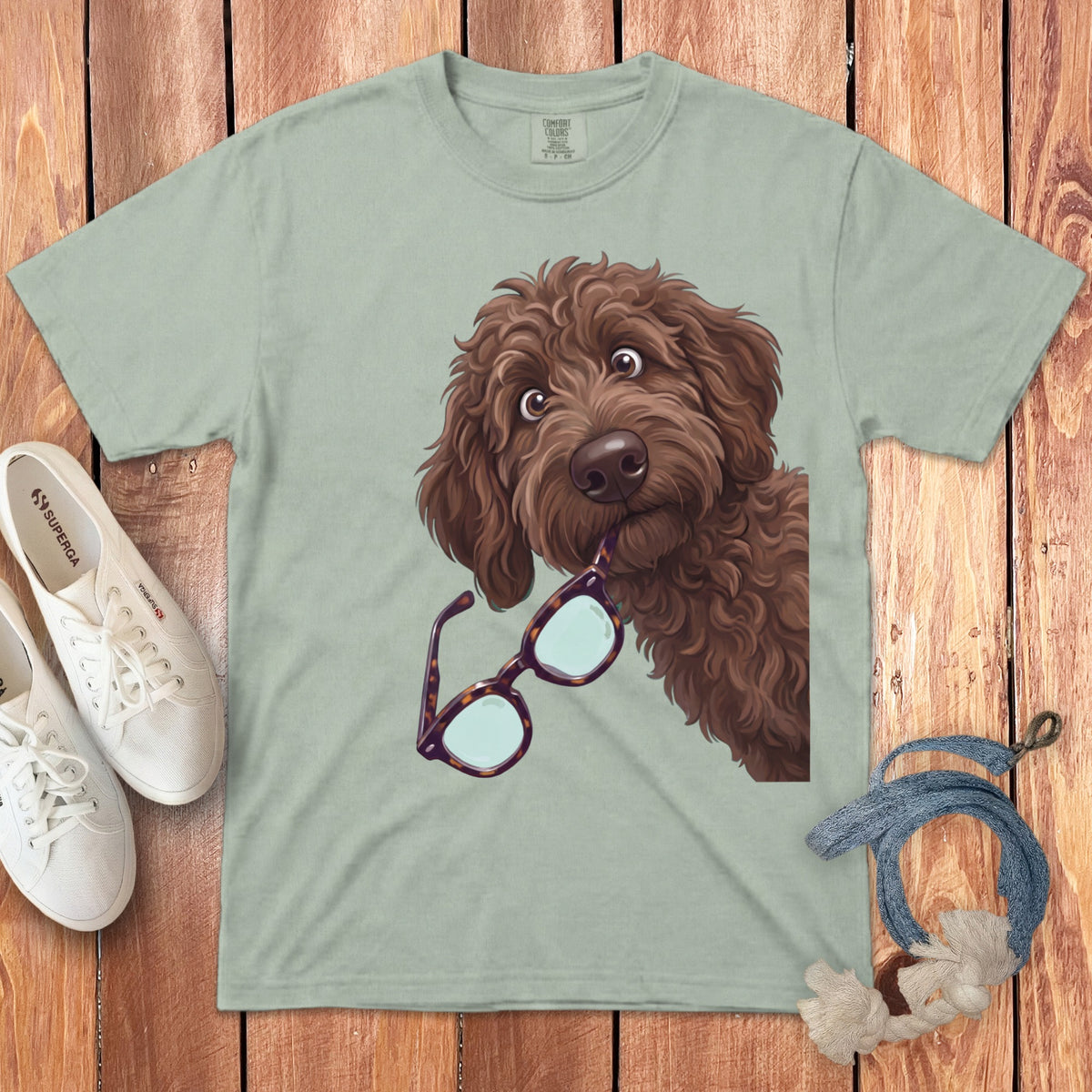 Sage green t-shirt featuring an illustration of a brown doodle dog holding glasses in its mouth, laid flat on wooden background.