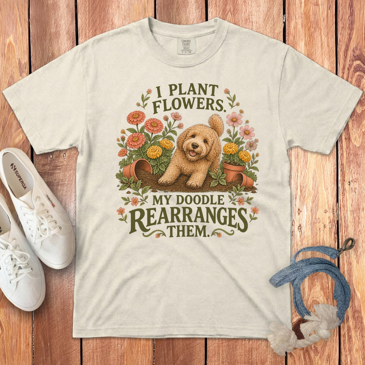 Beige crewneck t-shirt featuring a cartoon doodle dog in a garden with the text I plant flowers, my doodle rearranges them.