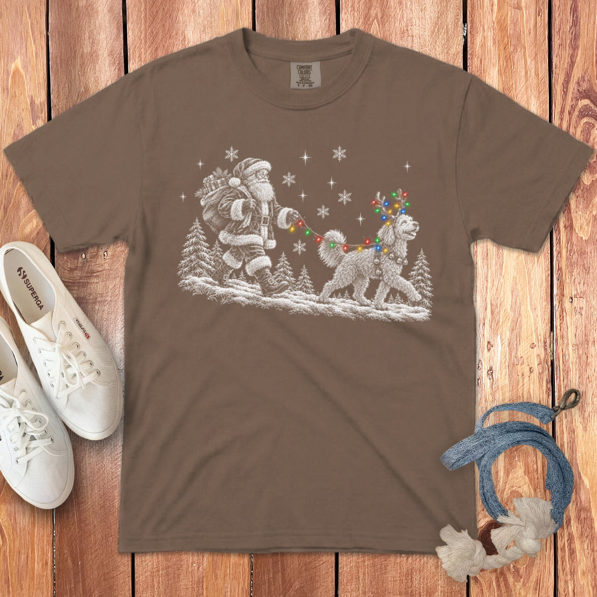 Brown t-shirt featuring a white graphic of Santa walking a dog with string lights, laid on wood with shoes and rope.
