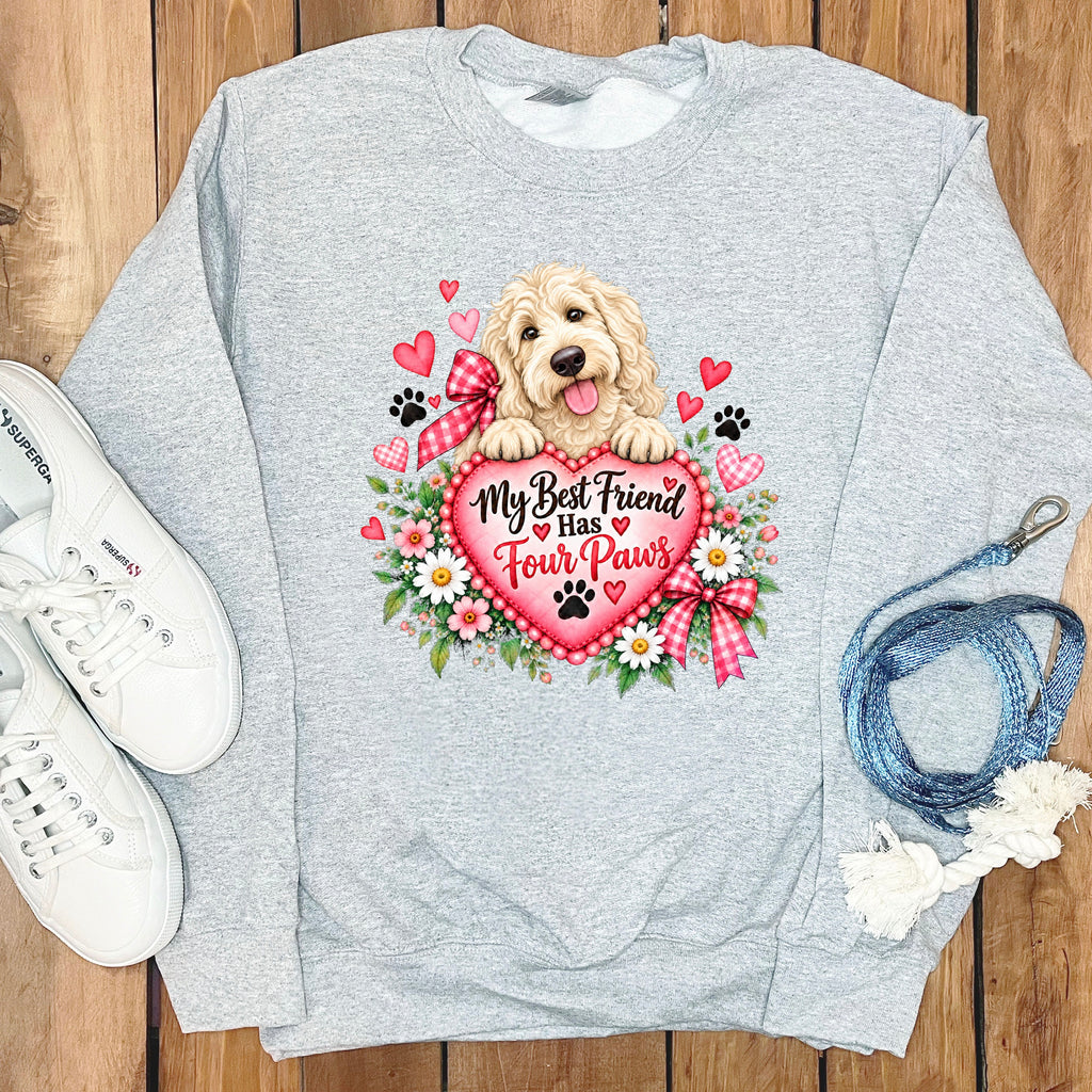 Heather gray crewneck sweatshirt featuring a graphic of a golden retriever holding a pink heart with the text My Best Friend Has Four Paws, surrounded by flowers, hearts, and paw prints, flat-laid on a wooden surface with white sneakers and a blue leash.