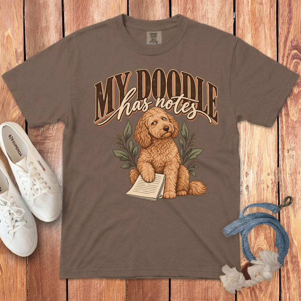 Brown t-shirt featuring a cartoon Goldendoodle and the text My Doodle has notes on a wooden background.