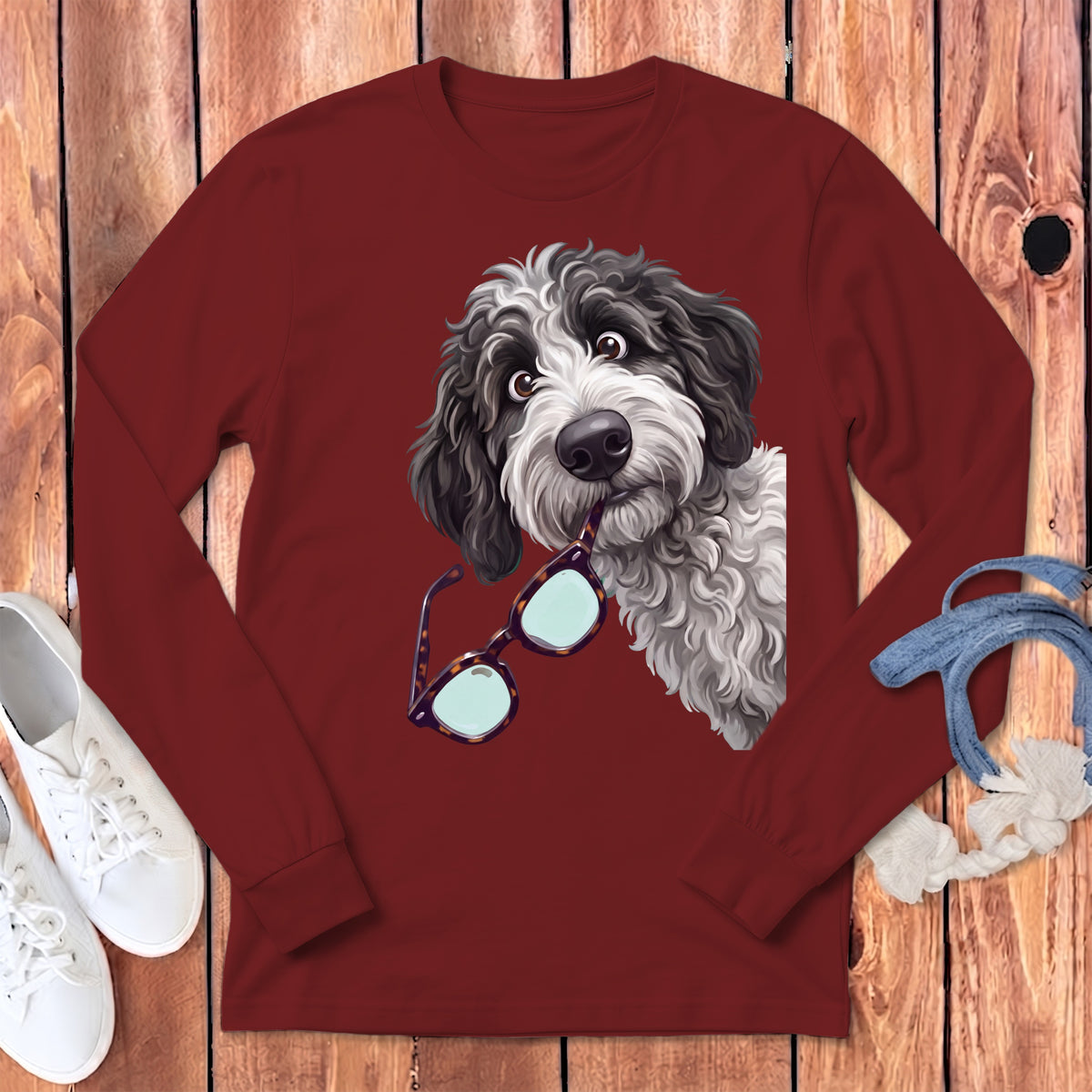 Maroon long-sleeve t-shirt featuring a cartoon dog holding glasses in its mouth, laid on a wooden background with white sneakers.