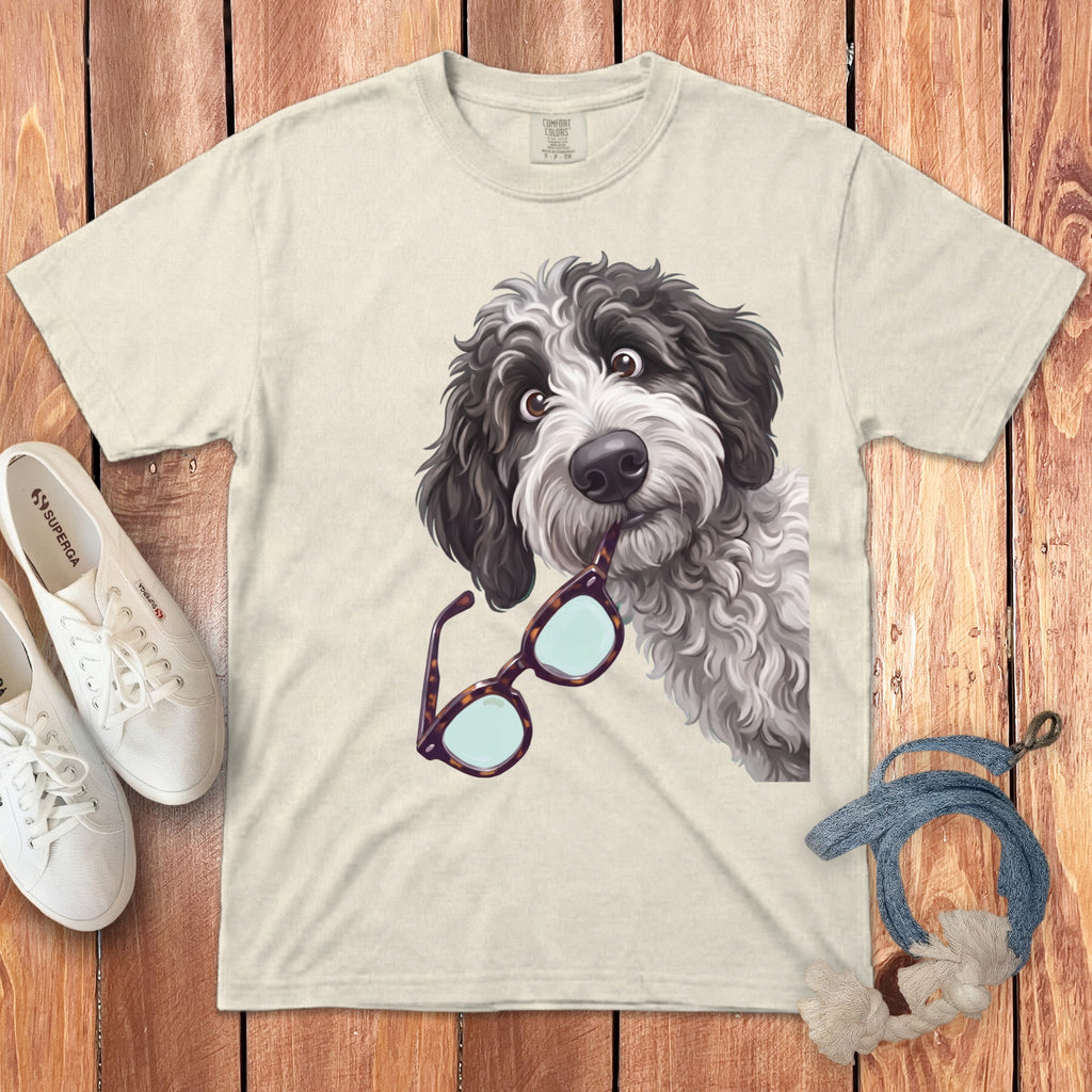 Beige t-shirt featuring a cartoon dog holding glasses in its mouth, laid flat on wooden planks.