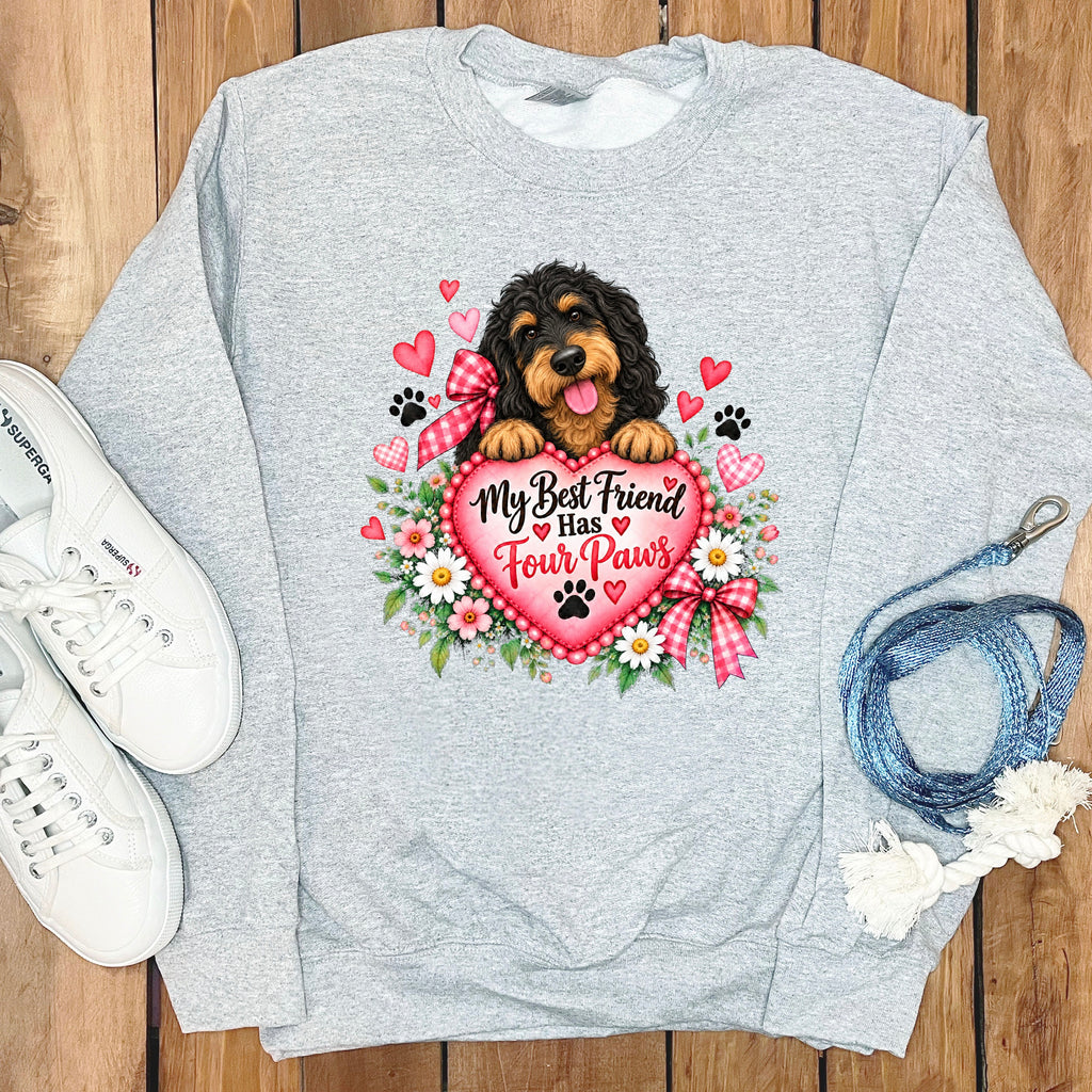 Light gray crewneck sweatshirt featuring a graphic of a black and tan doodle dog holding a large pink heart with the text My Best Friend Has Four Paws, surrounded by flowers, hearts, and bows, laid out on a wooden surface with white sneakers and a blue leash.