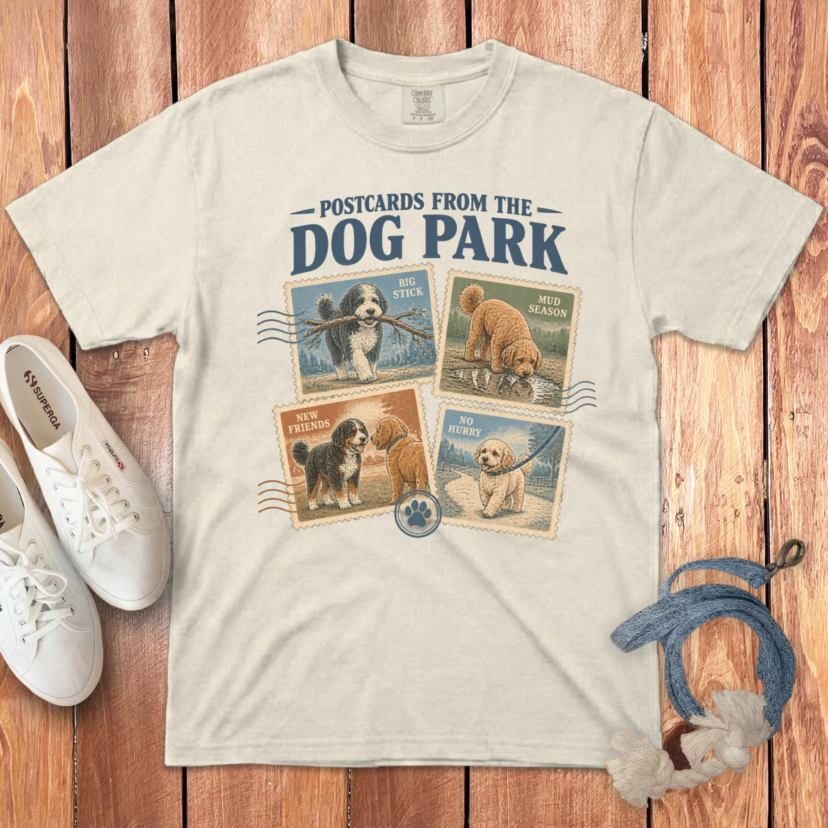 Cream-colored t-shirt featuring a dog park graphic with four postage stamp designs, surrounded by sneakers and a leash.