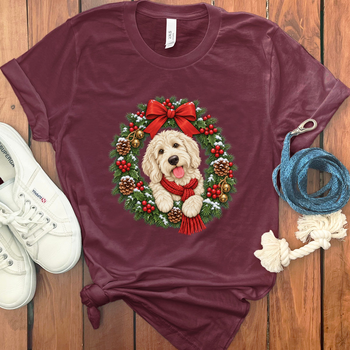 Maroon t-shirt featuring a Golden Retriever puppy inside a Christmas wreath, accompanied by white sneakers and a blue leash on a wooden background.