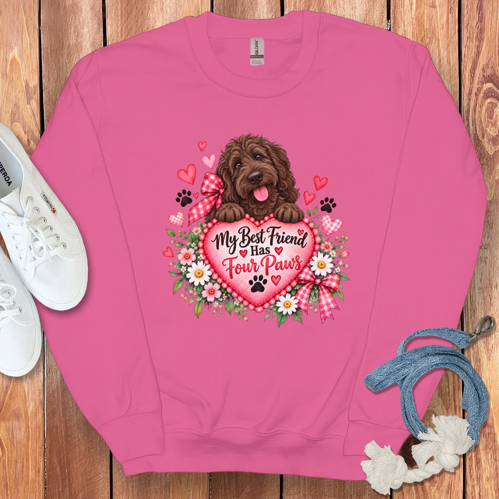 Pink crewneck sweatshirt featuring a graphic of a brown doodle dog resting on a heart with daisies and the text My Best Friend Has Four Paws, accompanied by white sneakers and a blue leash on a wooden background.
