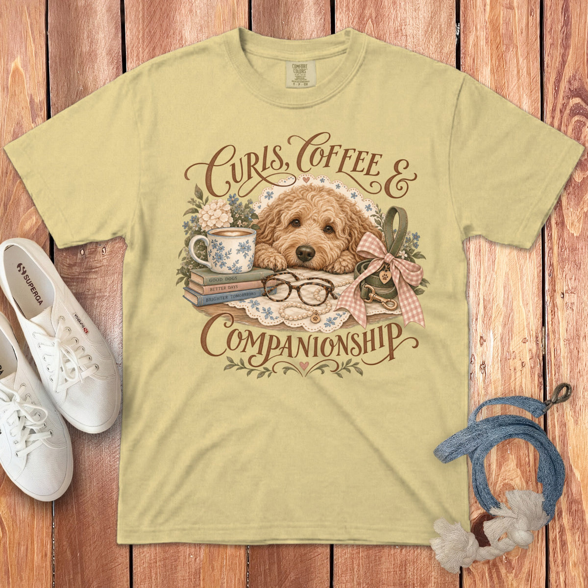 A pale yellow t-shirt featuring a vintage illustration of a fluffy golden doodle dog, books, and coffee.