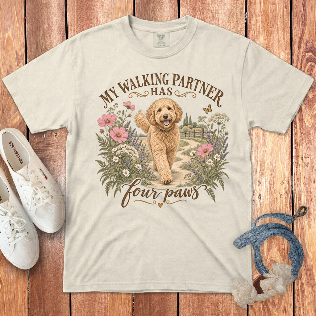 Cream t-shirt featuring a graphic of a golden doodle dog walking among flowers with text reading My walking partner has four paws.