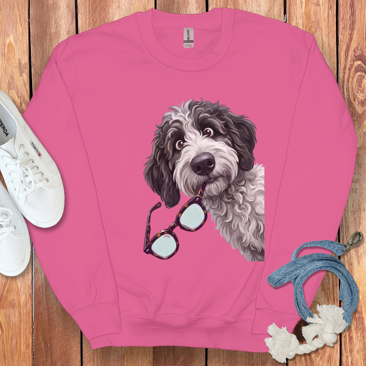 A pink crewneck sweatshirt featuring a graphic of a shaggy dog holding glasses in its mouth, laid flat on wood with white sneakers and a blue leash.