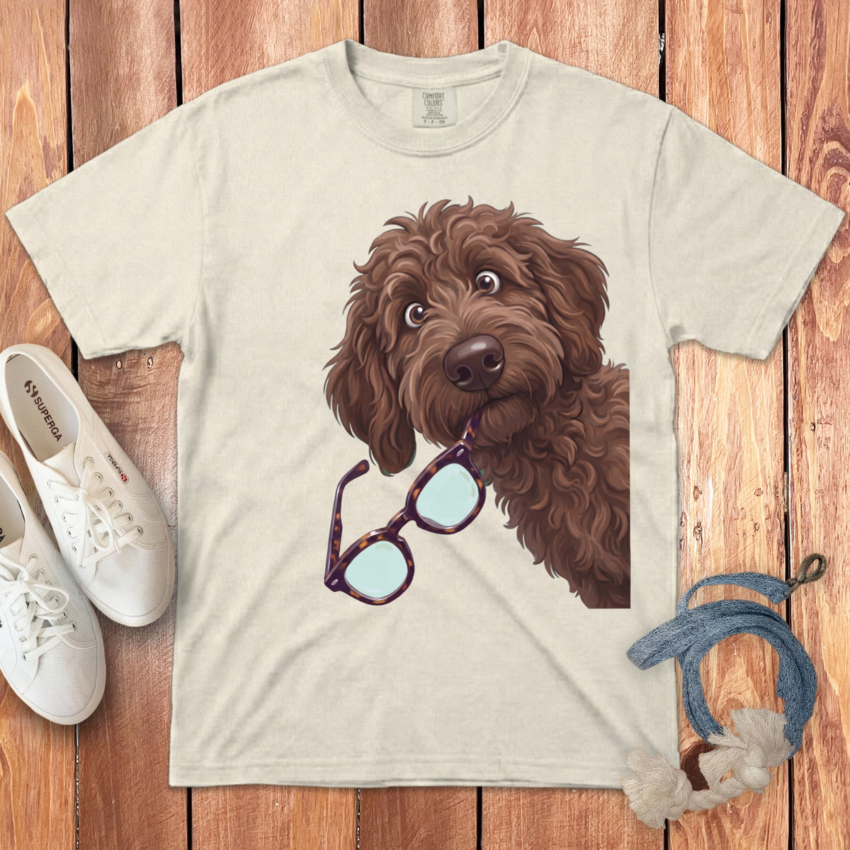 A cream-colored t-shirt featuring a cartoon illustration of a brown doodle dog holding glasses in its mouth.