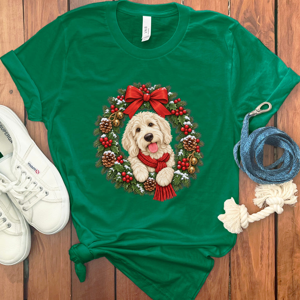 A kelly green t-shirt features a graphic of a fluffy golden doodle dog wearing a red scarf inside a Christmas wreath, laid out on a wooden surface with white sneakers and a blue leash.