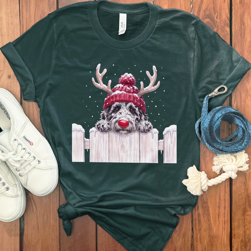 Dog wearing reindeer antlers and hat peeking over a snowy fence.