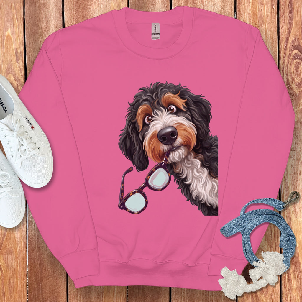 A pink crewneck sweatshirt features an illustrated Bernedoodle biting a pair of glasses on a wooden surface.