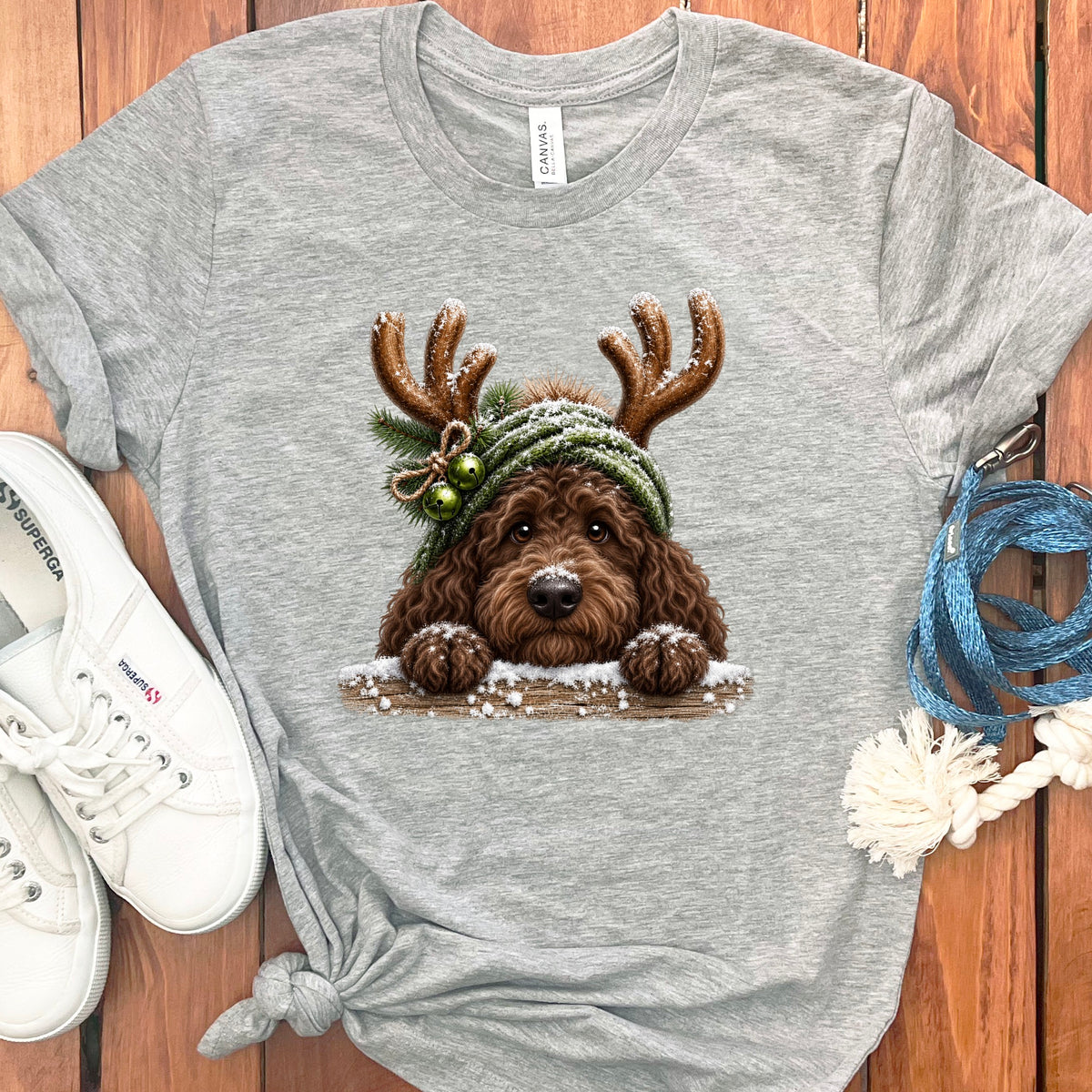Brown doodle dog wearing reindeer antlers and a green hat with bells, peeking over a snowy ledge.