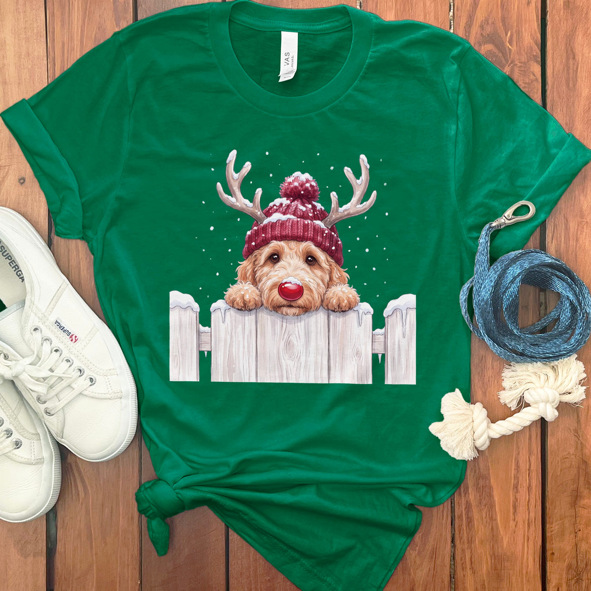 Green t-shirt with a doodle dog wearing a reindeer hat and red nose behind a white fence.