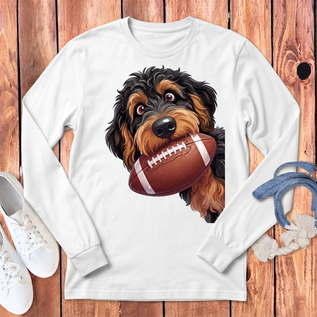 Fluffy black and brown dog holding a football in its mouth on white long-sleeve shirt.