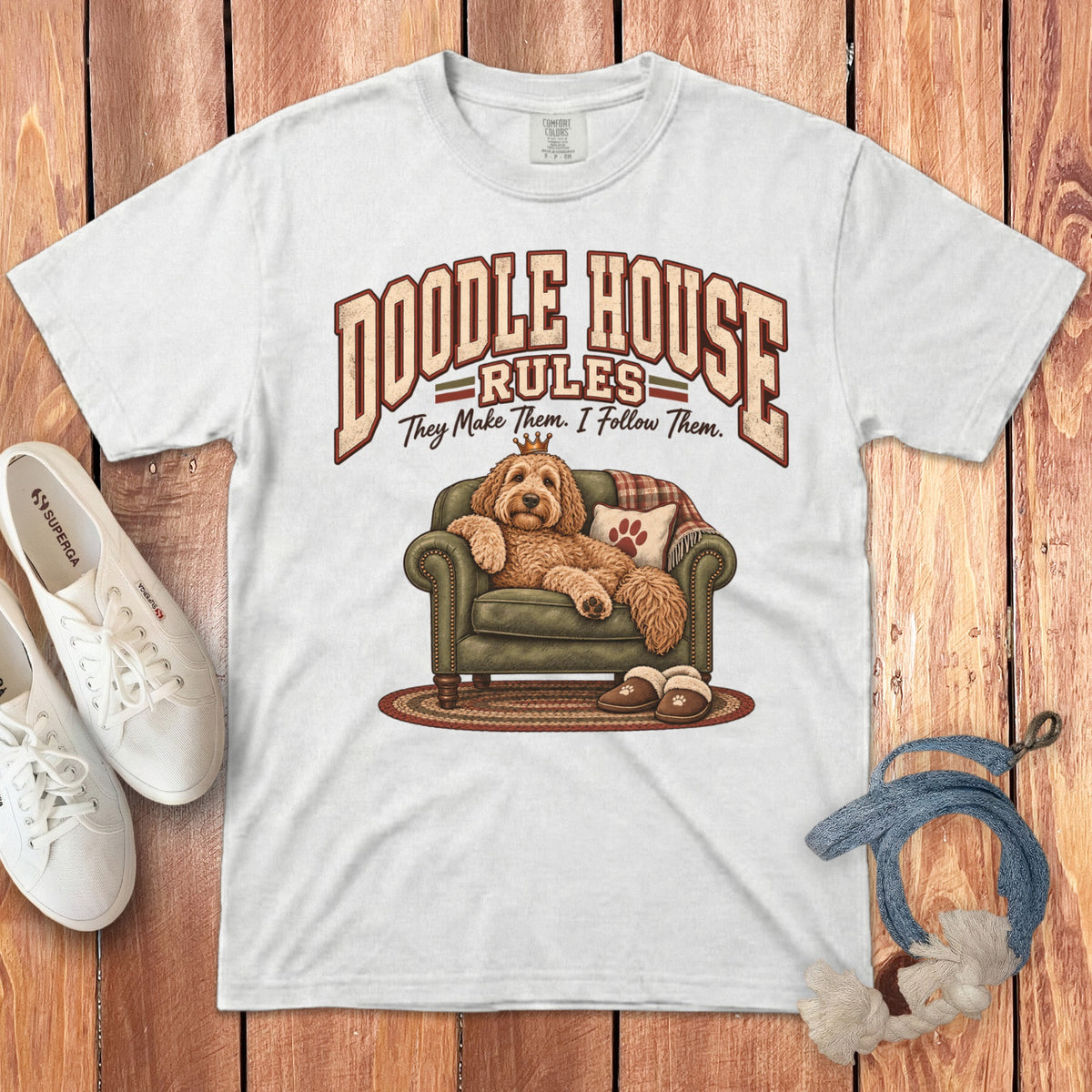 A white t-shirt featuring a graphic of a golden doodle wearing a crown and lounging on a green armchair.